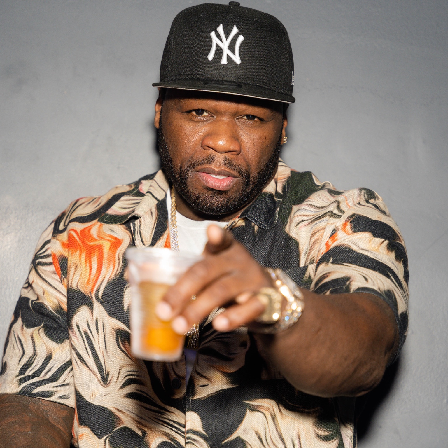A guest in a black baseball cap and patterned shirt raises a cup and gestures toward the camera.