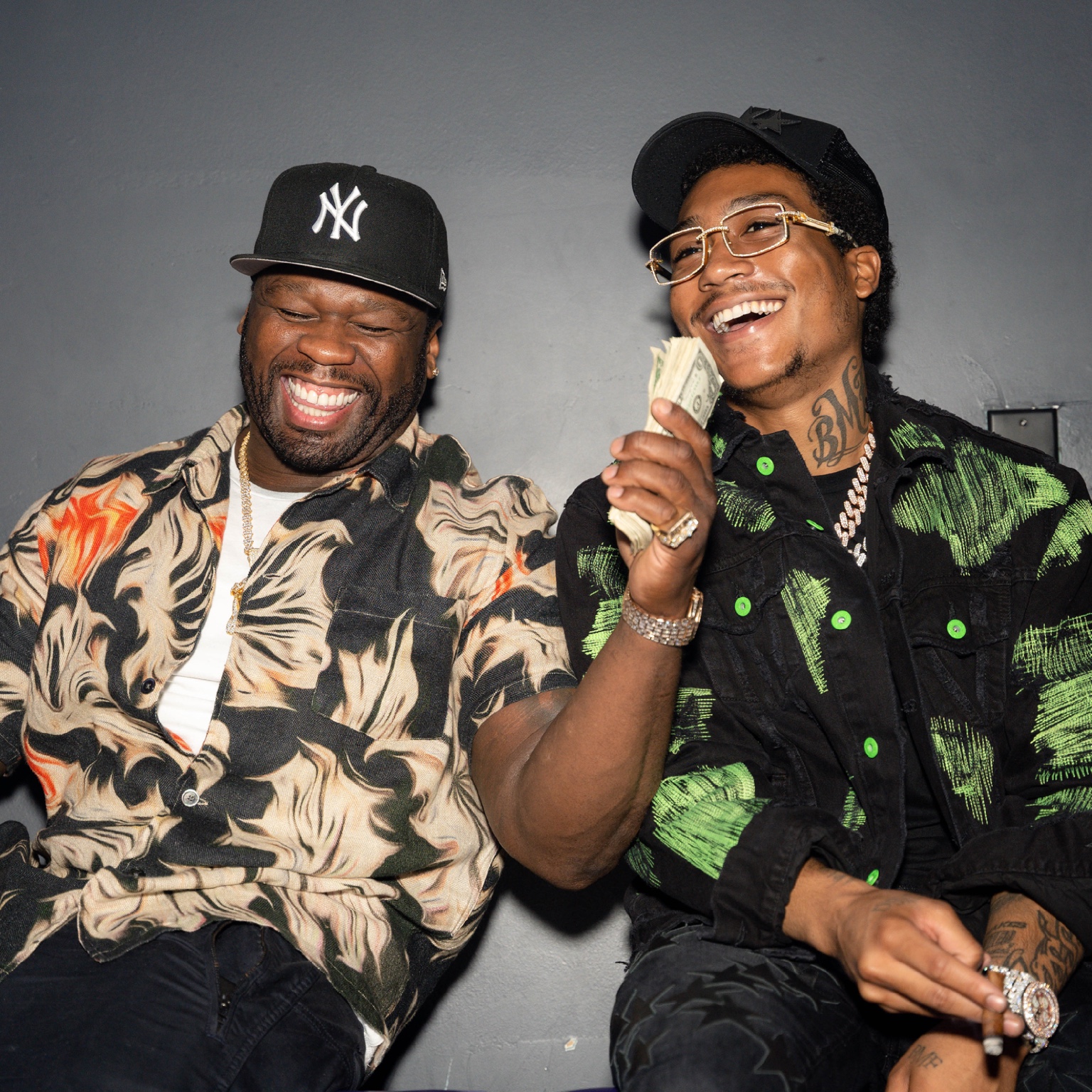 Two seated guests laughing together in patterned shirts and black caps during an indoor gathering.