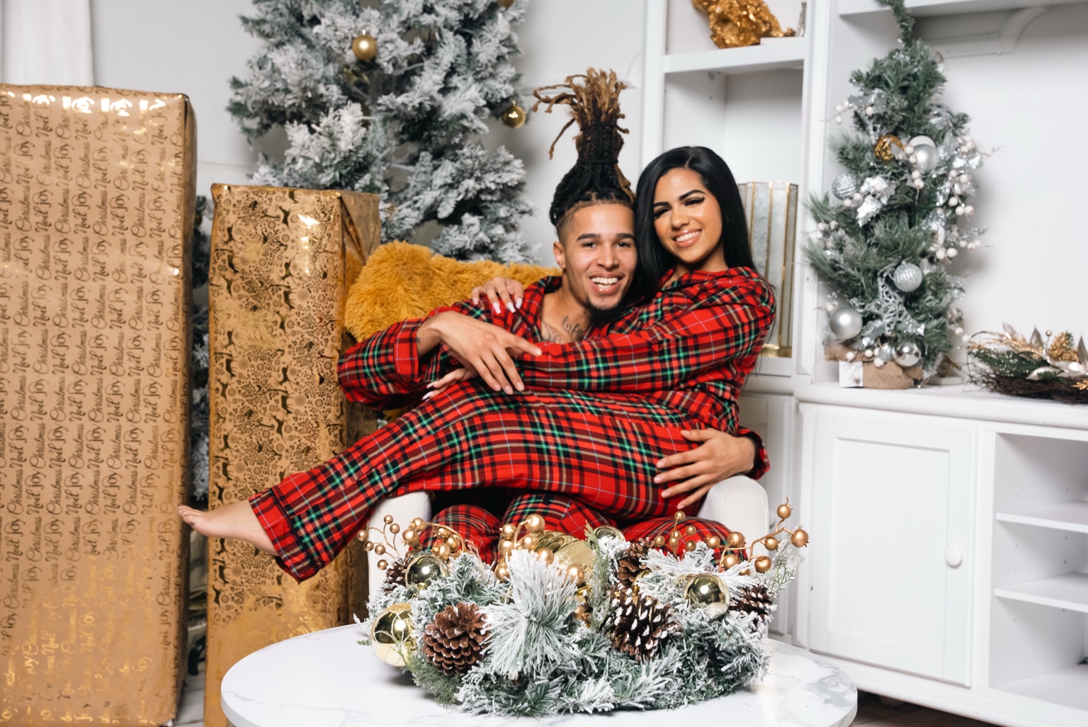 A smiling couple in matching red plaid pajamas seated together among snowy Christmas trees and gold wrapped gifts.
