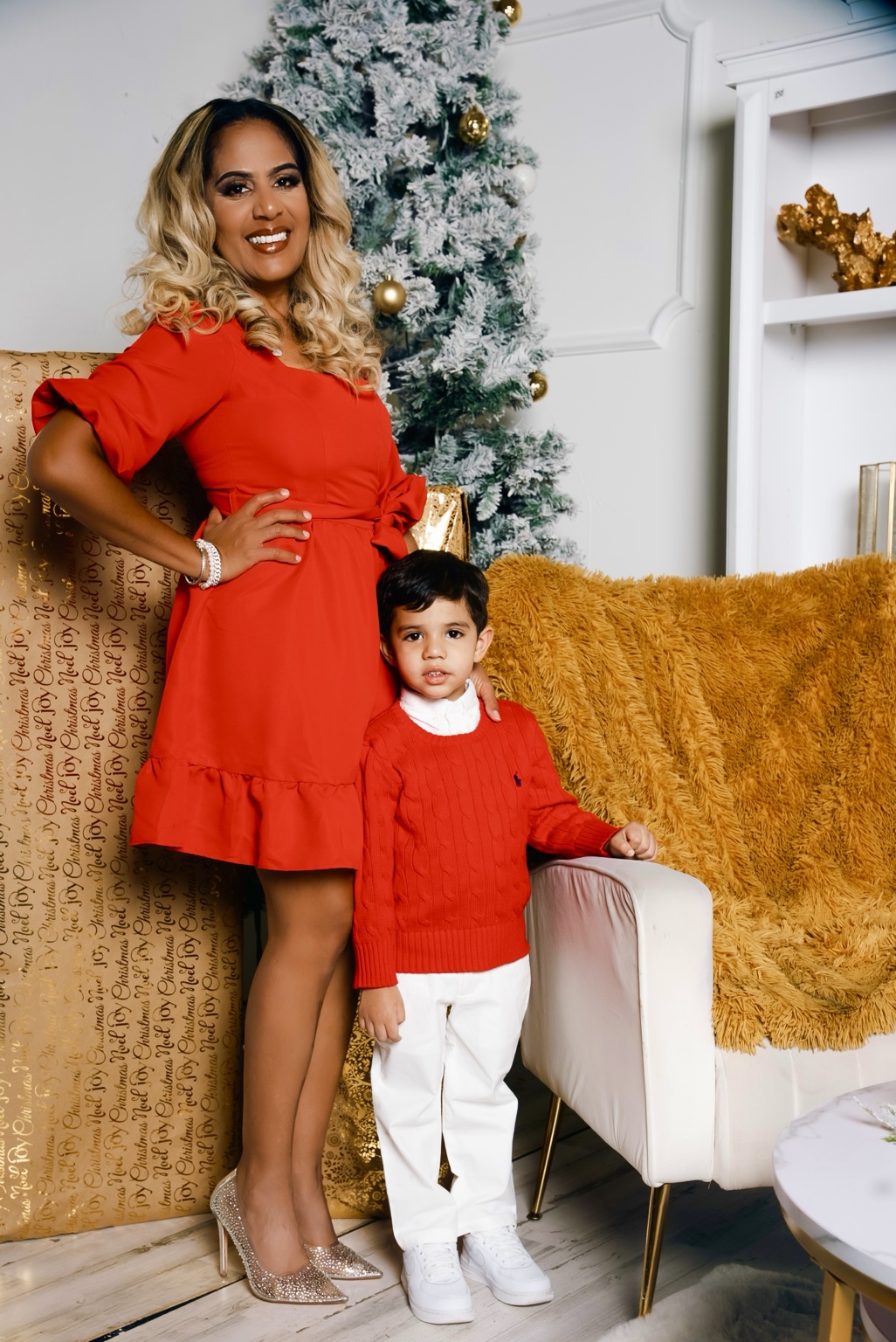An adult and child in coordinated red outfits beside a snowy Christmas tree, gold gifts, and a white chair.