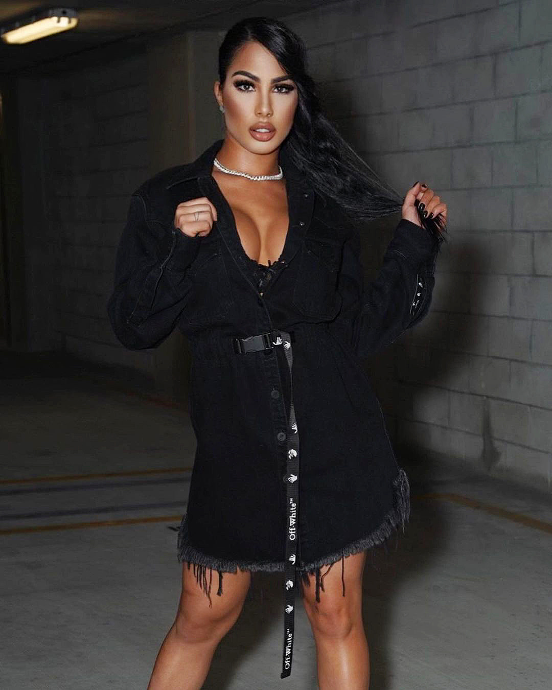 A portrait in a belted black denim outfit against the concrete walls of a parking garage.
