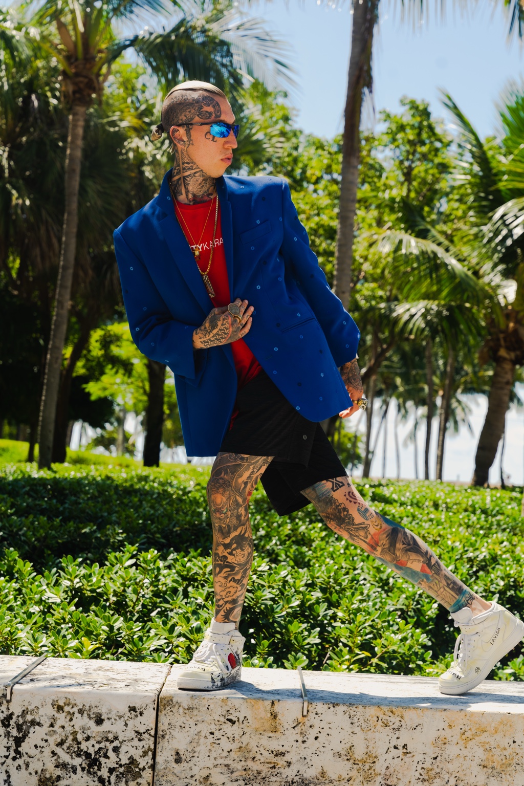 An outdoor fashion portrait in a blue blazer, red shirt, black shorts, and reflective sunglasses among palm trees.