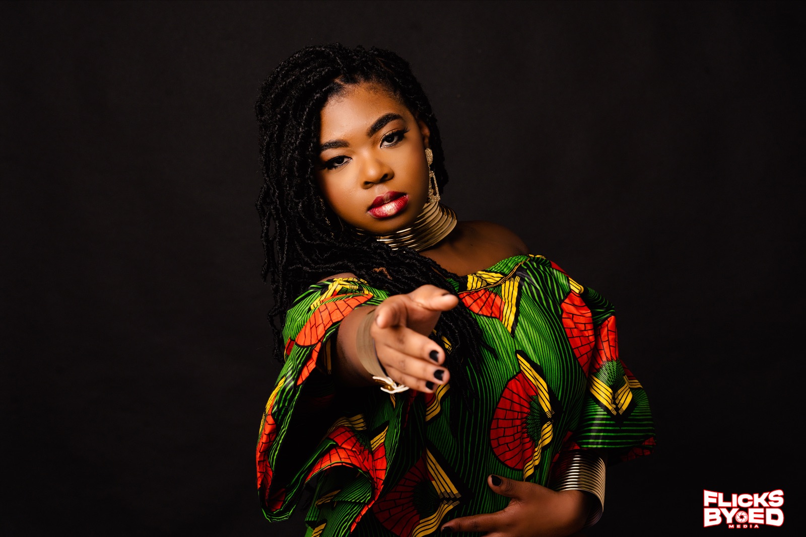A studio portrait with a vibrant green, red, and yellow patterned outfit, gold jewelry, and an outstretched hand against a dark backdrop.