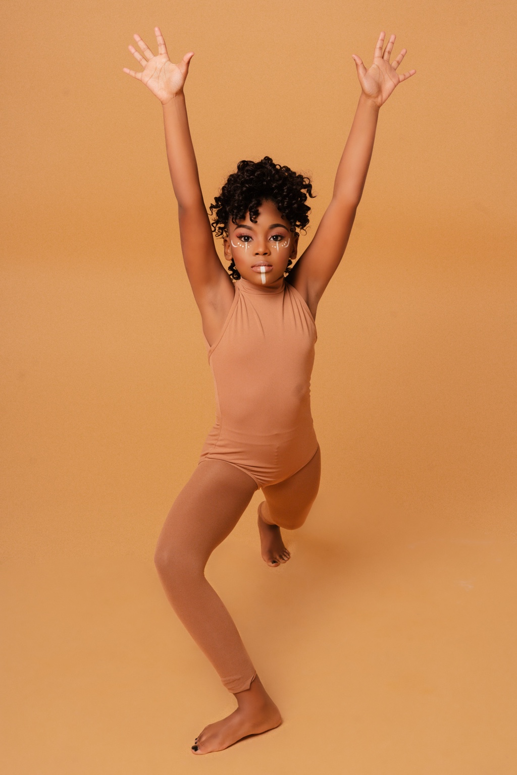 A young dancer raises both arms in a tan dance outfit against a warm gold studio backdrop.