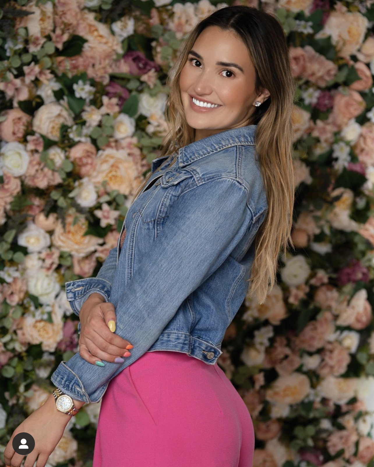 A smiling portrait in a denim jacket and pink outfit in front of a wall of pastel flowers.