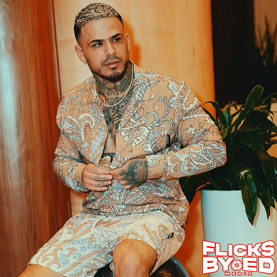 A seated fashion portrait in a coordinated patterned shirt and shorts beside a green plant.