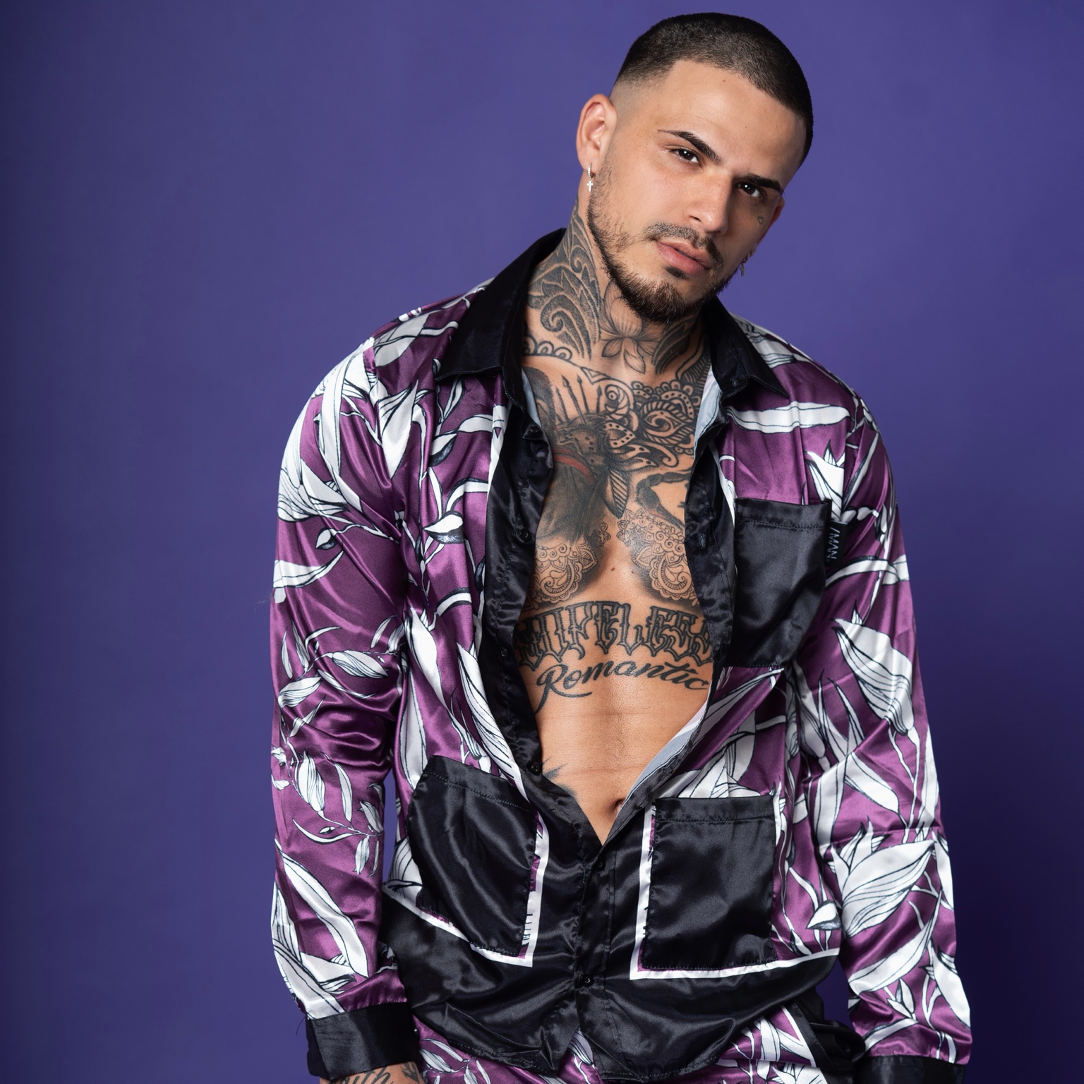 A studio portrait of a tattooed man in a patterned purple shirt against a purple backdrop.