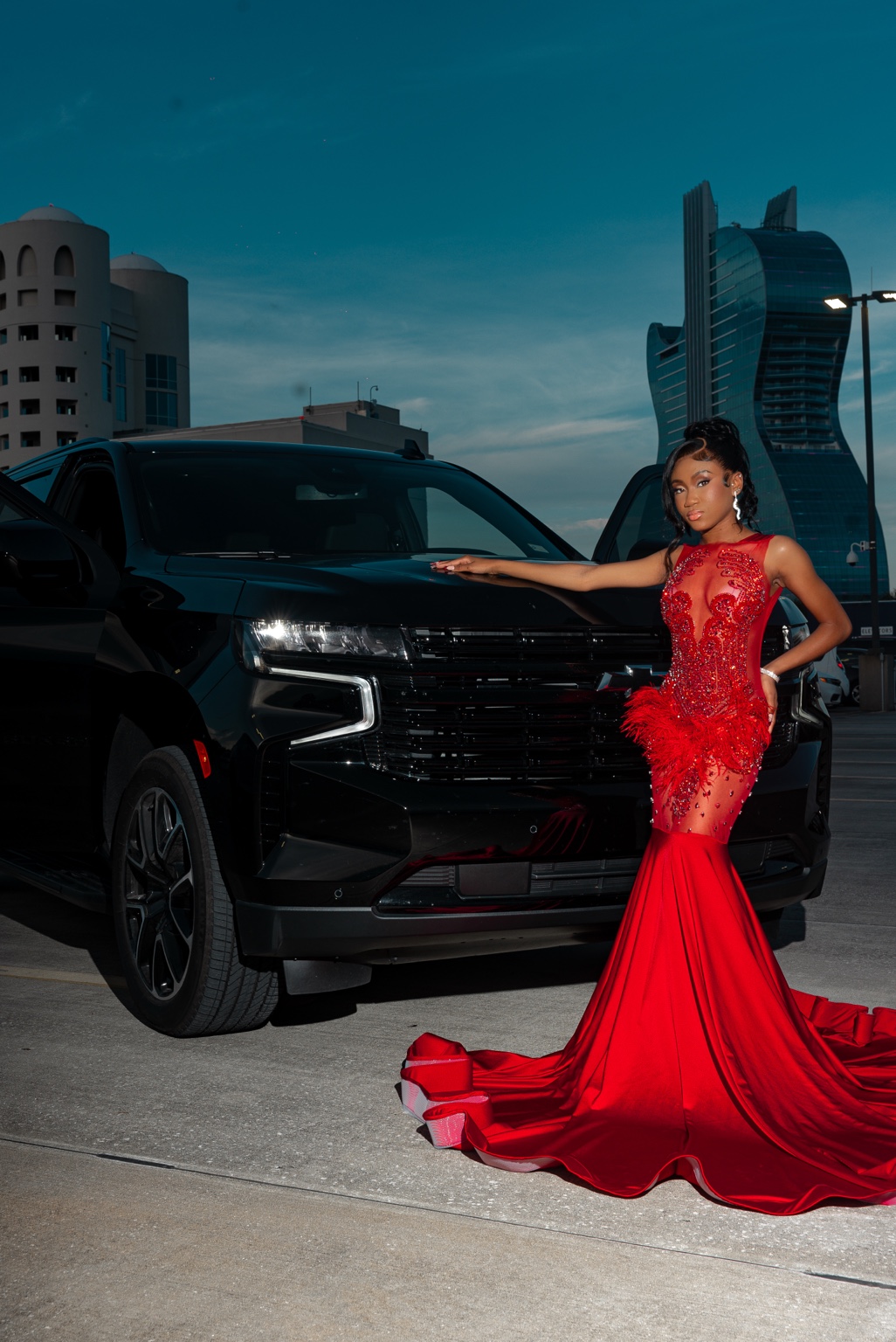 A full-length portrait in a red embellished gown beside a black SUV, with the guitar-shaped hotel in the background.